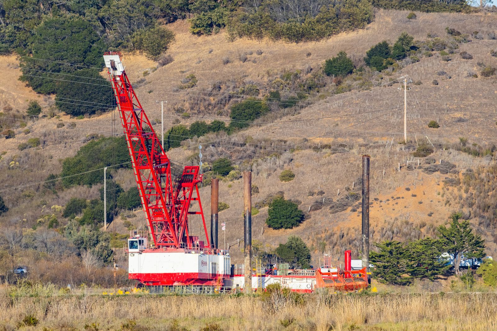 A red and white lattice-boom crawler crane working beside tall steel piles below dry golden coastal hills