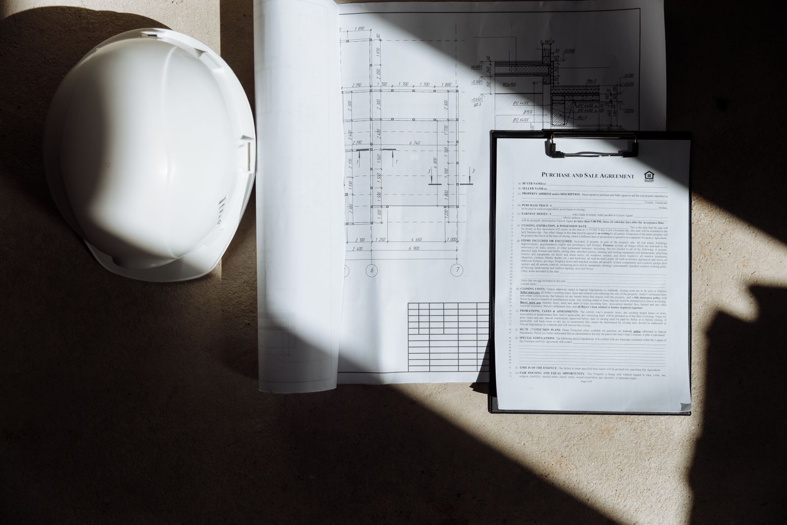 A white hard hat beside printed plans and a clipped inspection document lying in sunlight
