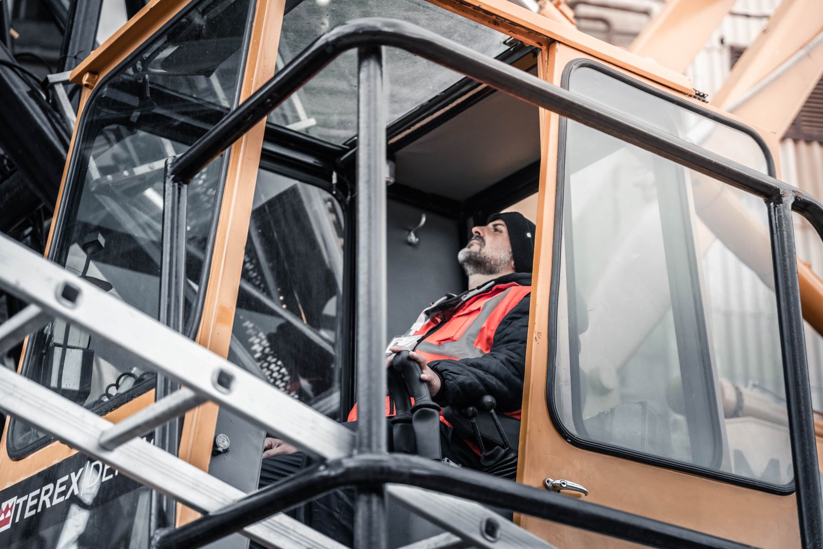 A crane operator in a hi-vis jacket and beanie seated at the controls inside an orange crane cab