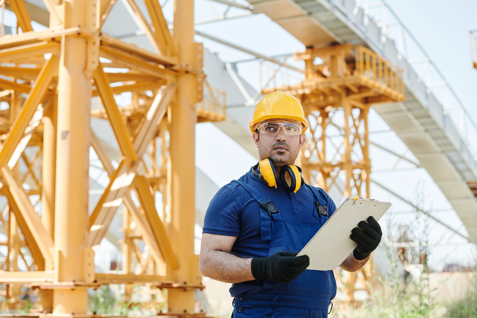 A crane inspector in a yellow hard hat, safety glasses and blue coveralls holding a clipboard beside yellow steel framework