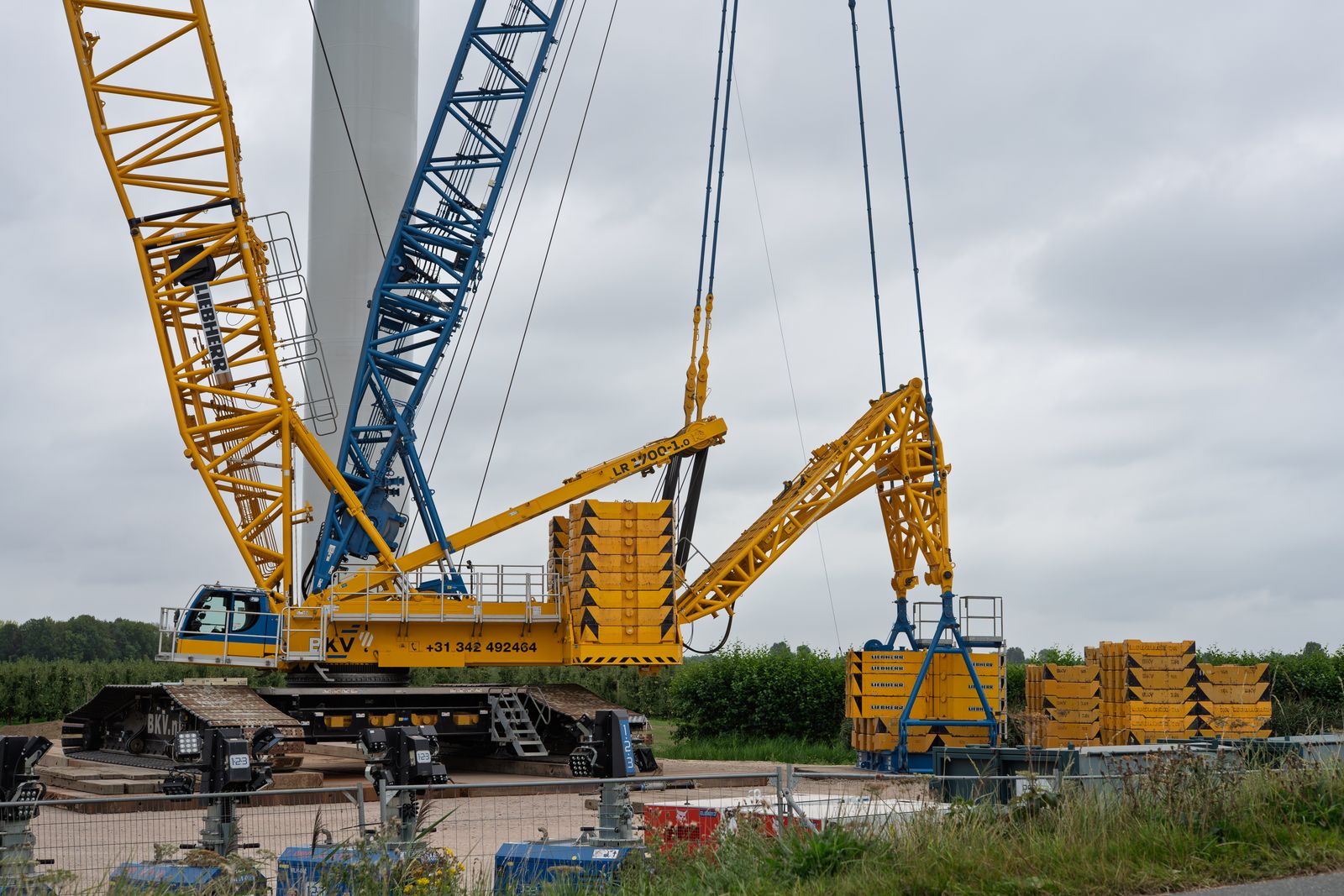 A yellow lattice-boom crawler crane rigged with counterweight on a fenced job site under an overcast sky