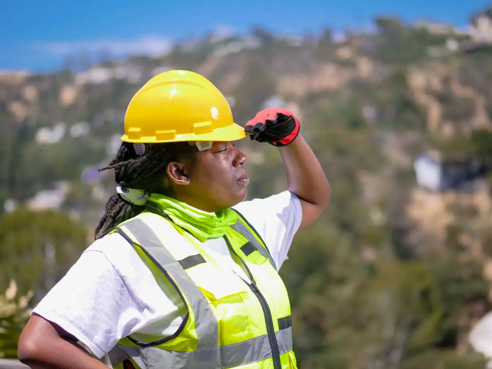 A construction worker in a yellow hard hat and high-visibility vest shading her eyes while watching work overhead outdoors.